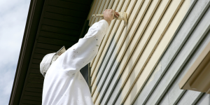 Man in white coveralls and hat paints exterior of house light yellow.