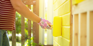 Female homeowner in flower-printed glove paints exterior of home with yellow paint on sunny day.