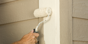 Homeowner uses paint roller to paint trim on exterior of home white.
